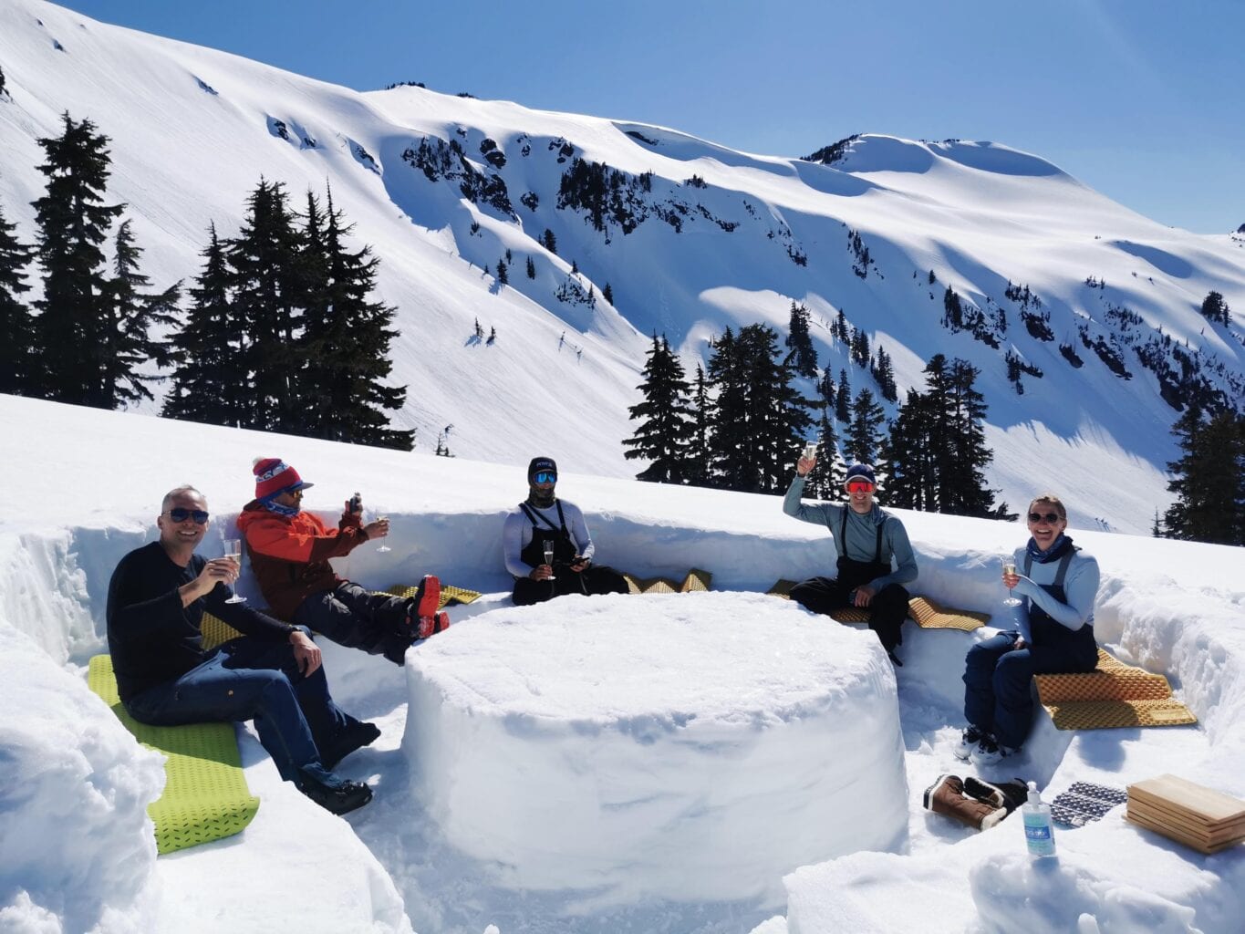 Mabey Ski guests sit around snow table with glass of champagne in the sun. Whistler, BC
