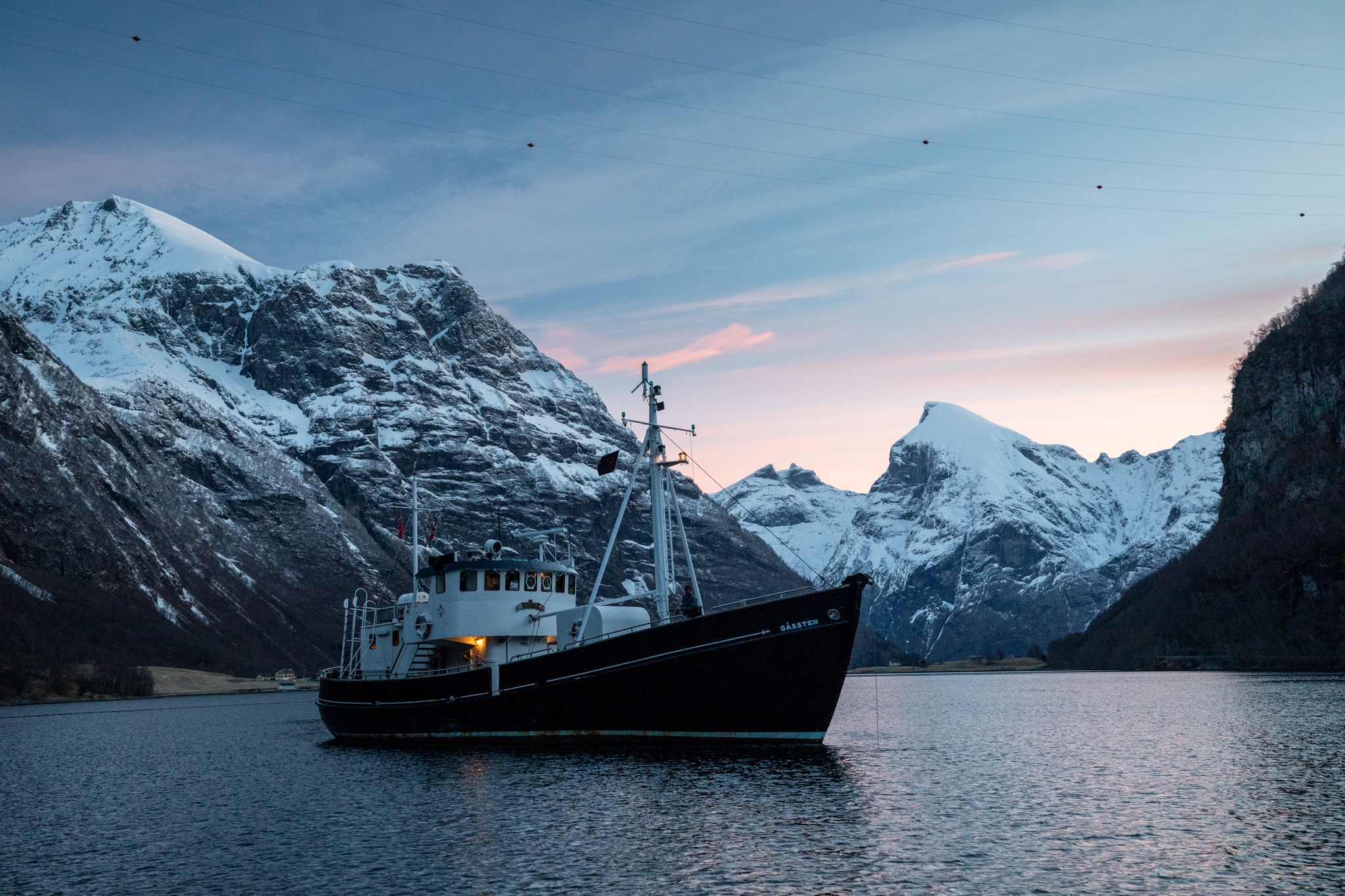 HMS Gassten boat floats in a Norwegian fjord with snowy mountains behind