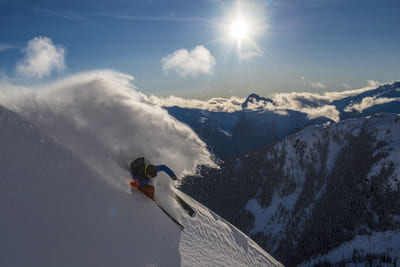 skier going down hill in powder snow with mountains and sun in the background